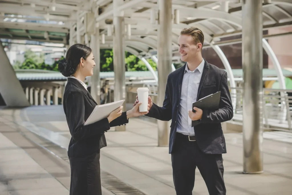 Businesswoman and businessman exchange a coffee cup and smile in a bright, modern transit station, conveying a friendly professional meeting.