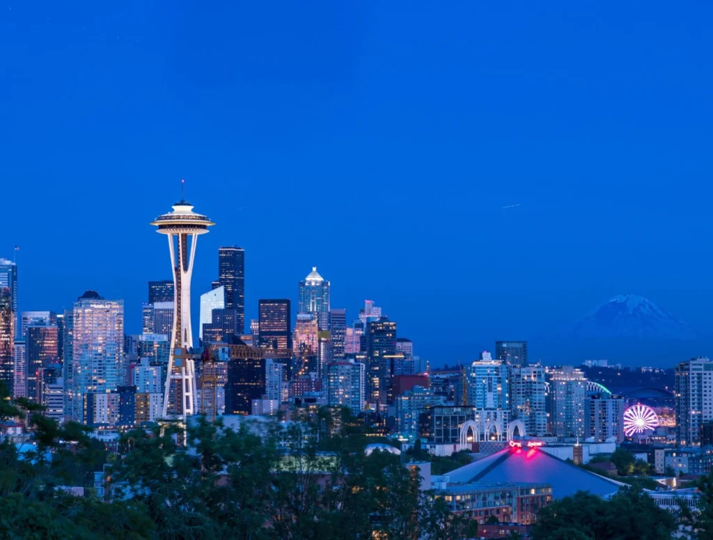 Twilight view of Seattle skyline with the illuminated Space Needle, Mount Rainier, city lights, and a glowing Ferris wheel under blue skies.