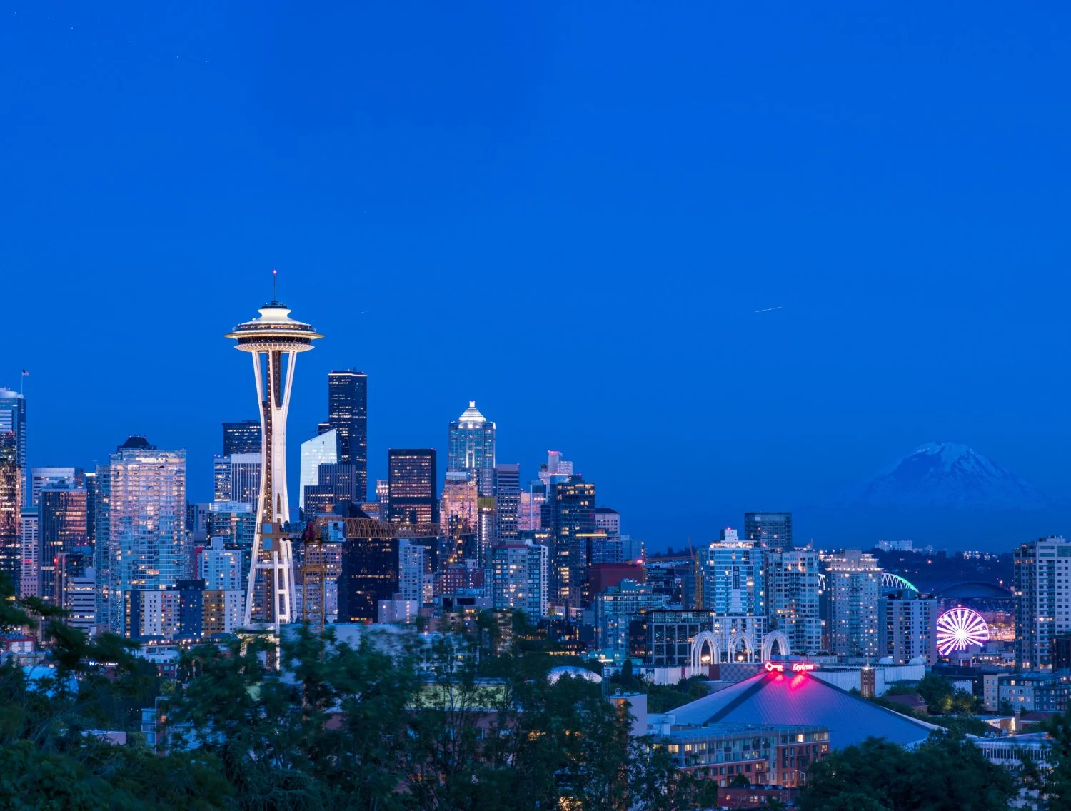 Twilight view of Seattle skyline with the illuminated Space Needle, Mount Rainier, city lights, and a glowing Ferris wheel under blue skies.