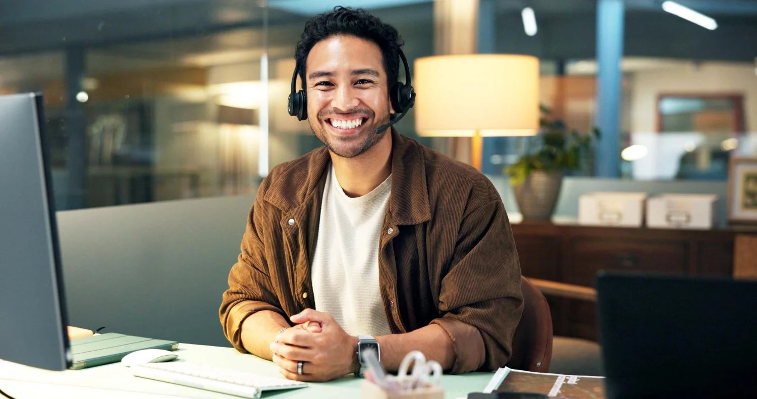 A smiling man wearing a headset sitting at a desk with a computer and office supplies.