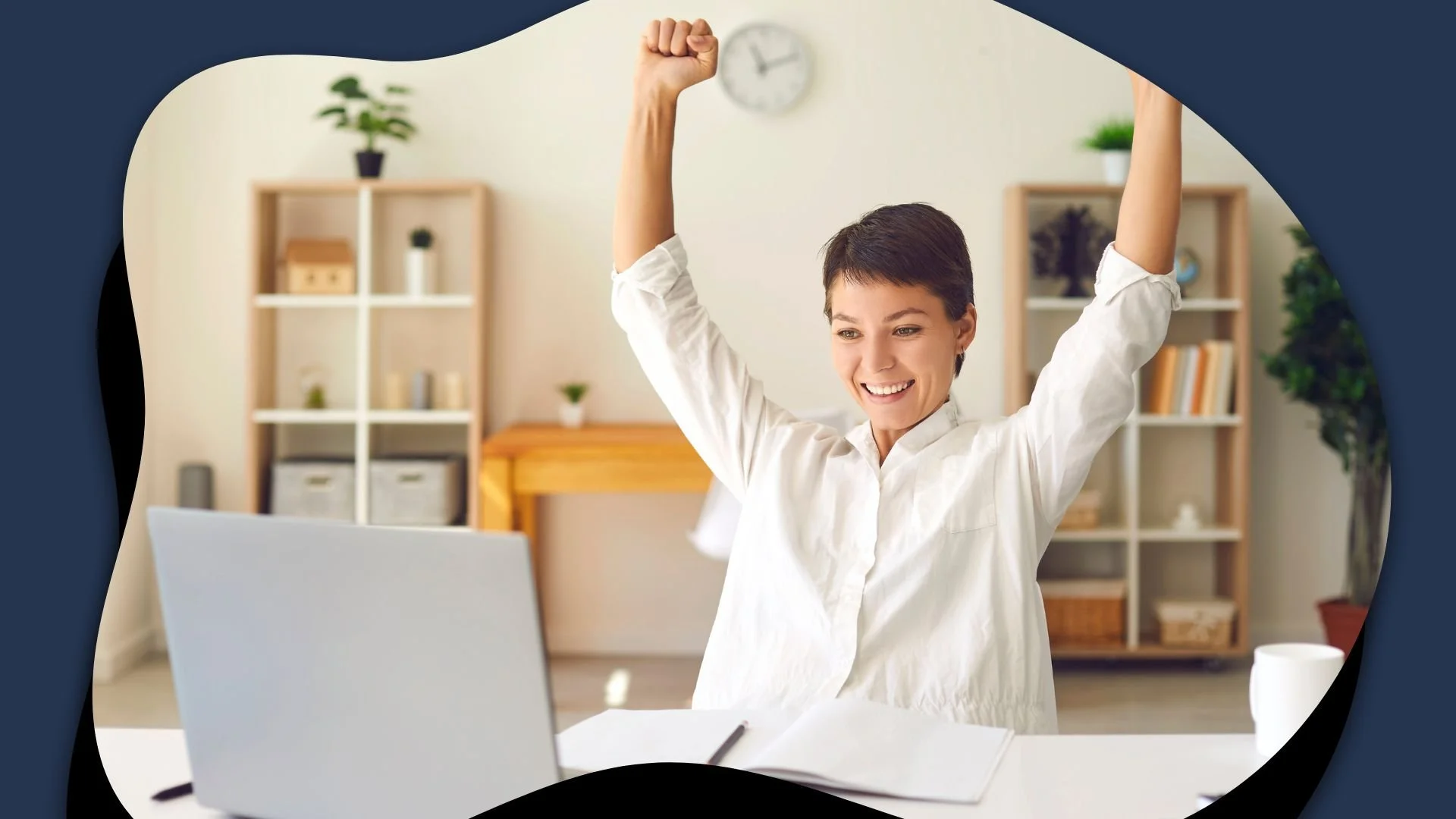 Smiling woman in a white blouse raises both arms triumphantly behind a laptop in a home office, conveying workplace success and confidence.