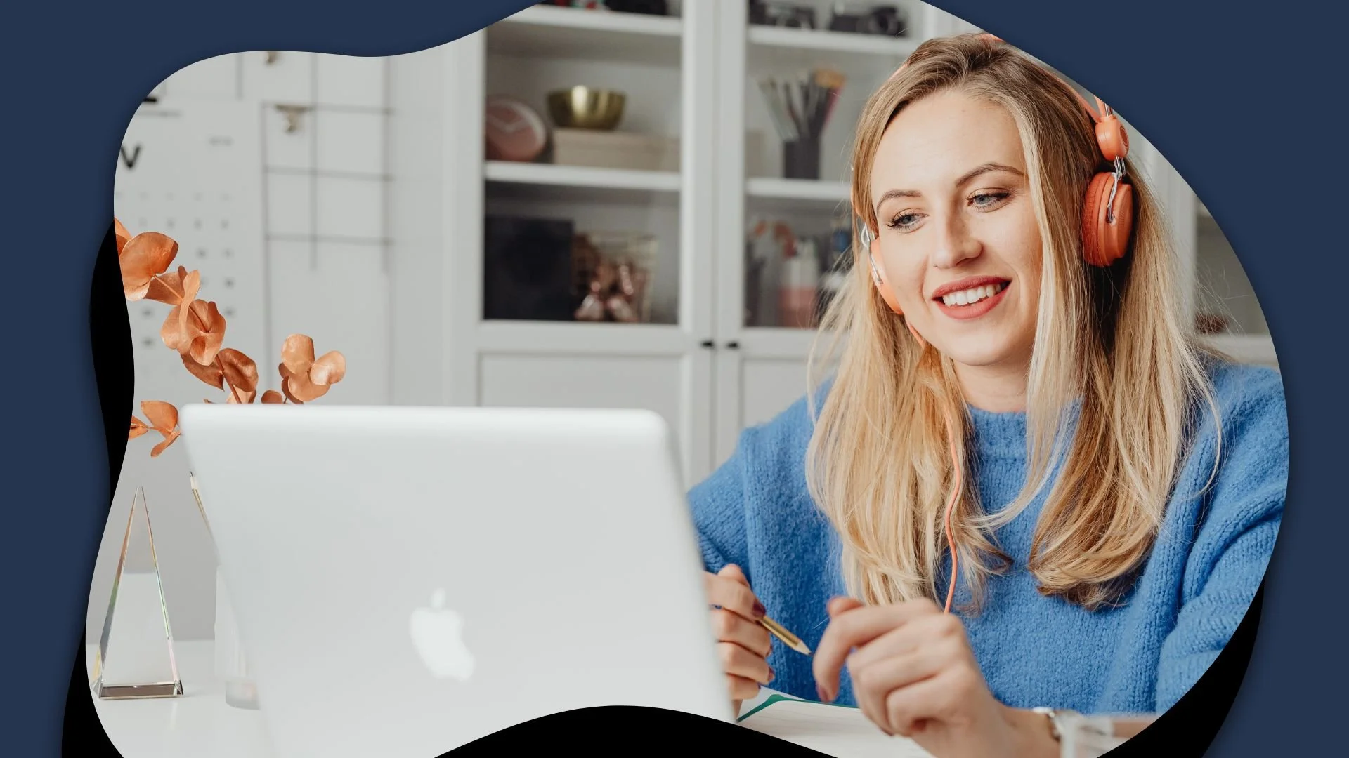 Smiling blonde woman in a blue sweater and orange headphones works on a laptop at home, creating a cheerful, focused atmosphere.
